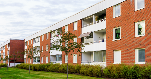 Modern three-story apartment building with red brick and white paneling, featuring balconies and a landscaped lawn with trees and shrubs.