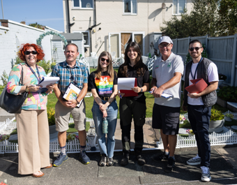 A group of six people stand outside in a sunny front garden, holding clipboards and notes. The garden features neatly maintained lawns, decorative borders and white edging, with a house and garden shed in the background. The group appears to be inspecting or judging the garden as part of a community gardening activity. Bright sunshine casts strong shadows across the garden and paved area.