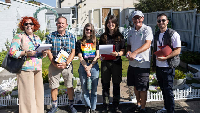 A group of six people stand outside in a sunny front garden, holding clipboards and notes. The garden features neatly maintained lawns, decorative borders and white edging, with a house and garden shed in the background. The group appears to be inspecting or judging the garden as part of a community gardening activity. Bright sunshine casts strong shadows across the garden and paved area.