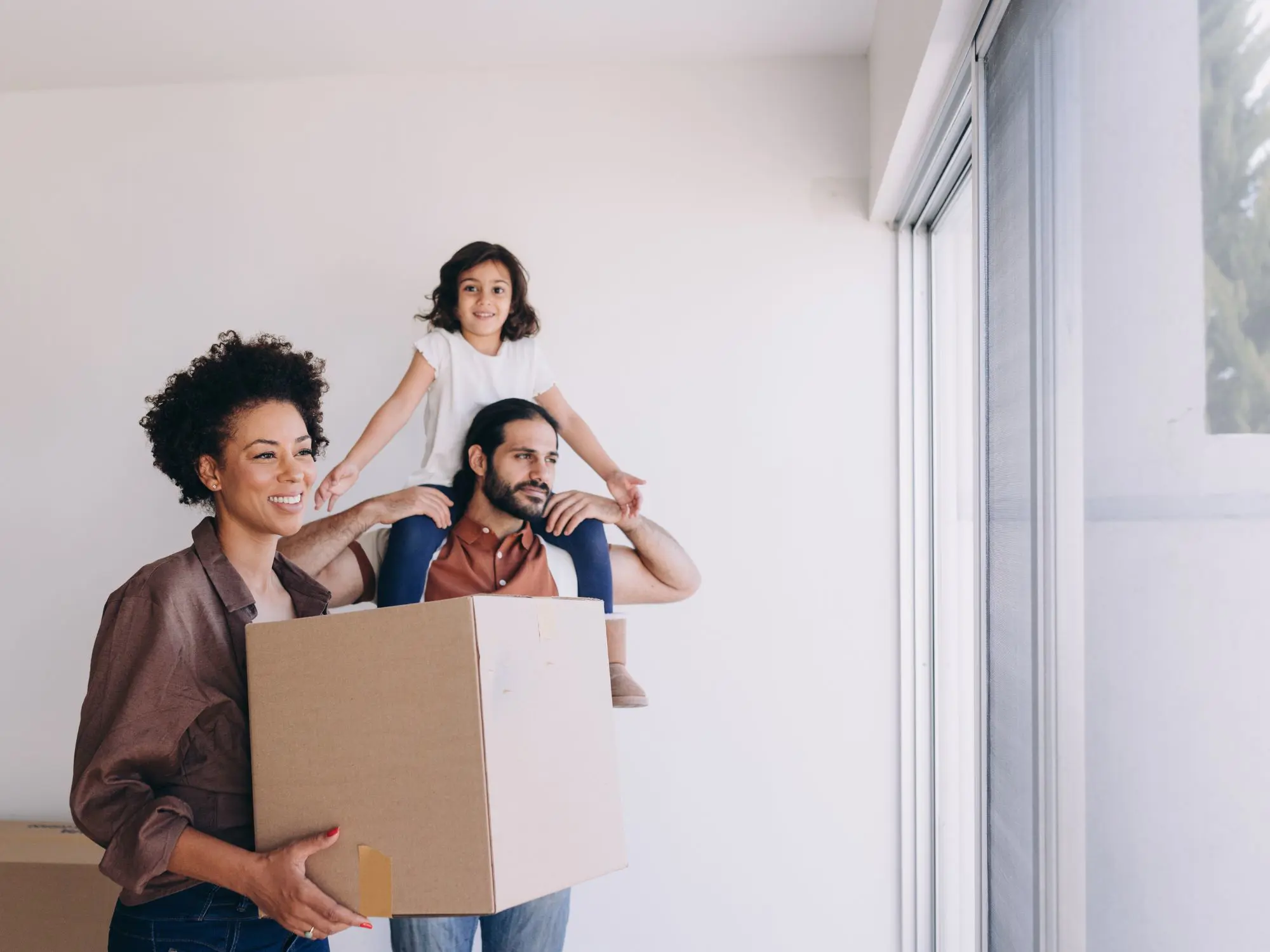 Three people in a bright room with white walls and a large window. One person holds a cardboard box, another carries a child on their shoulders with arms outstretched, suggesting a joyful moment during moving or unpacking.