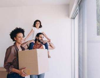 Three people in a bright room with white walls and a large window. One person holds a cardboard box, another carries a child on their shoulders with arms outstretched, suggesting a joyful moment during moving or unpacking.