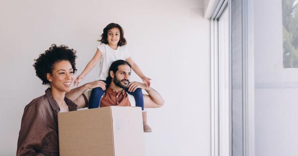 Three people in a bright room with white walls and a large window. One person holds a cardboard box, another carries a child on their shoulders with arms outstretched, suggesting a joyful moment during moving or unpacking.