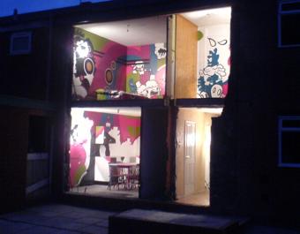 Interior view of a two-story building with the front wall removed, revealing brightly lit rooms decorated with colorful, cartoon-style murals. The upper room features green and pink abstract designs, while the lower room has pink and white artwork and a dining table with chairs. The scene is set against a dark evening sky.