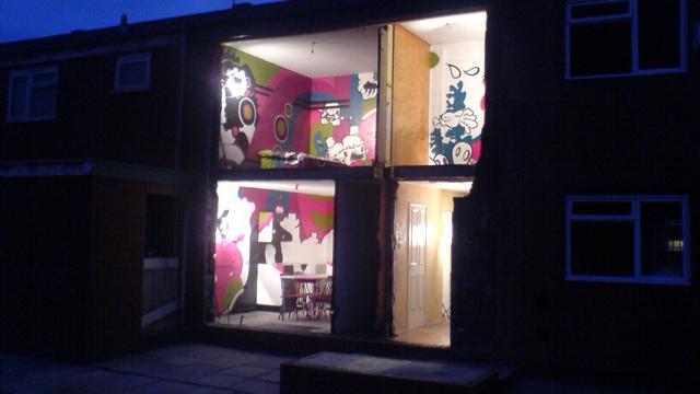 Interior view of a two-story building with the front wall removed, revealing brightly lit rooms decorated with colorful, cartoon-style murals. The upper room features green and pink abstract designs, while the lower room has pink and white artwork and a dining table with chairs. The scene is set against a dark evening sky.