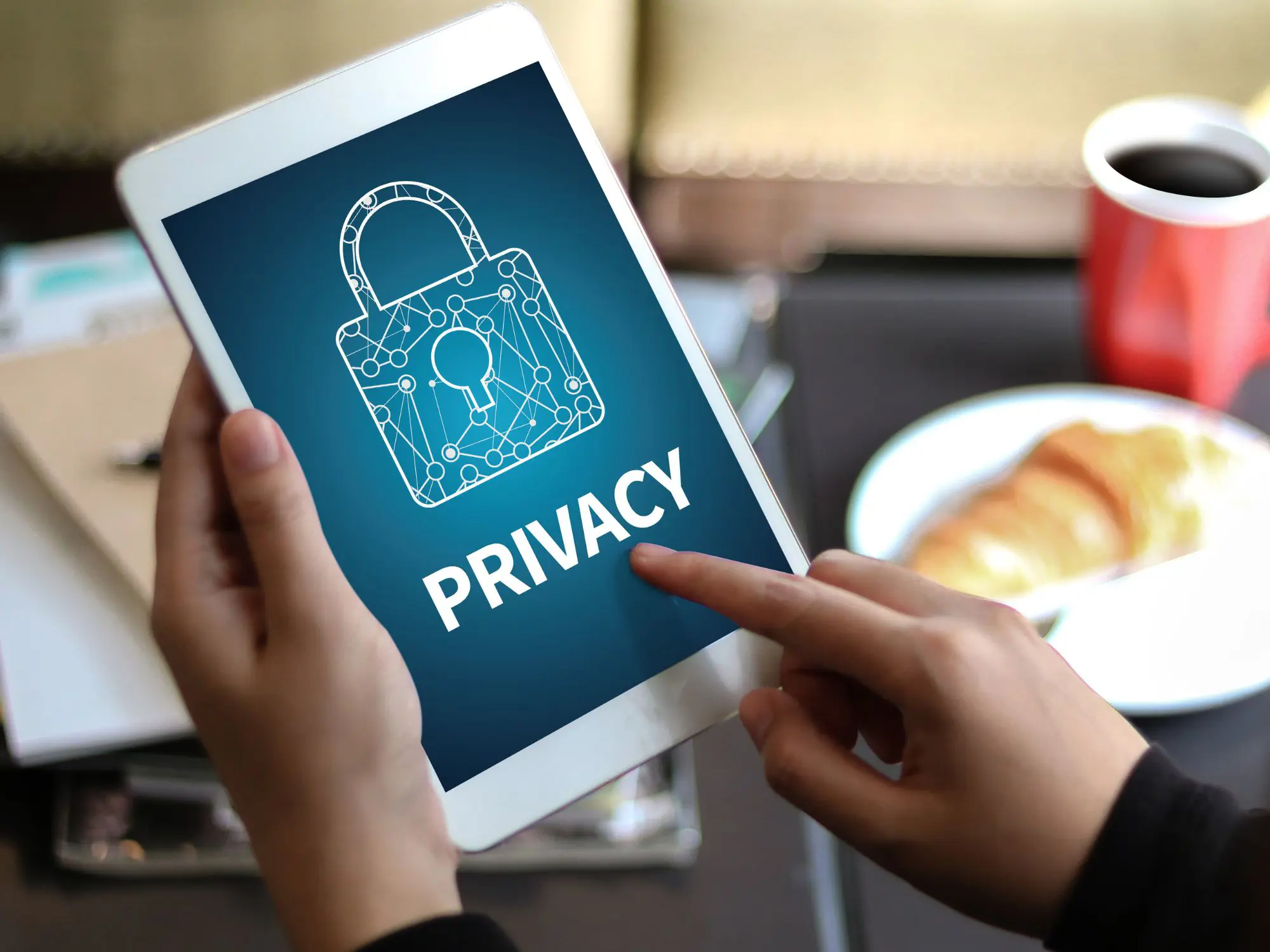 A person holding a tablet displaying an illustration of a padlock with a circuit-like design and the word "PRIVACY" below it. The background of the screen is blue. In the background, there are blurred objects including a cup of coffee and a plate with food.