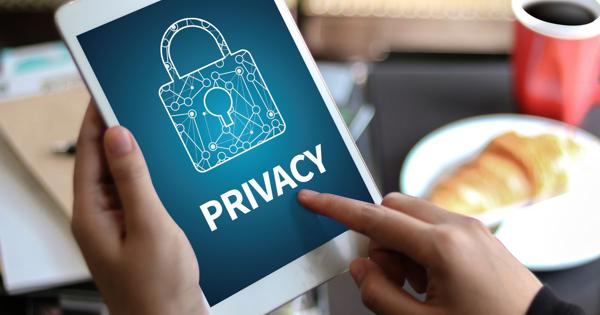 A person holding a tablet displaying an illustration of a padlock with a circuit-like design and the word "PRIVACY" below it. The background of the screen is blue. In the background, there are blurred objects including a cup of coffee and a plate with food.