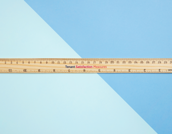 Wooden ruler with centimeter and inch markings placed diagonally on a two-tone blue background, featuring the text 'Tenant Satisfaction Measures'.