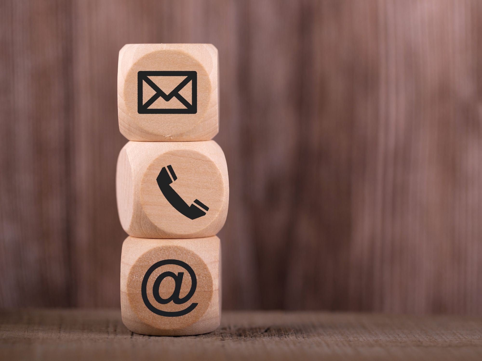 Stacked wooden blocks with contact icons: envelope for email, telephone for phone, and @ symbol for online contact, on a blurred wooden background.