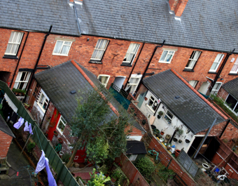 Row of red brick terraced houses with dark grey tiled roofs and fenced backyards, some featuring laundry, plants, and garden furniture.
