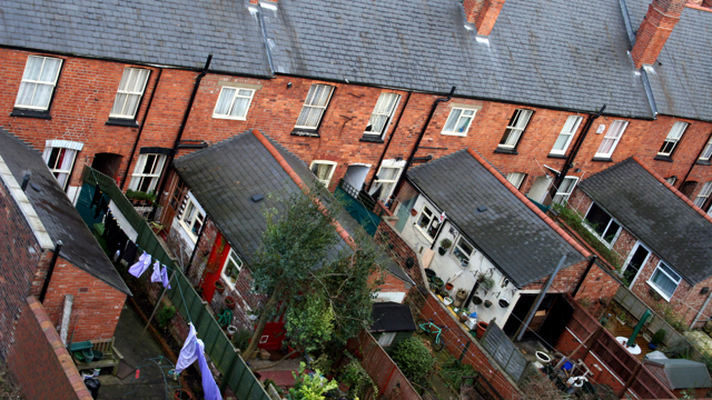Row of red brick terraced houses with dark grey tiled roofs and fenced backyards, some featuring laundry, plants, and garden furniture.