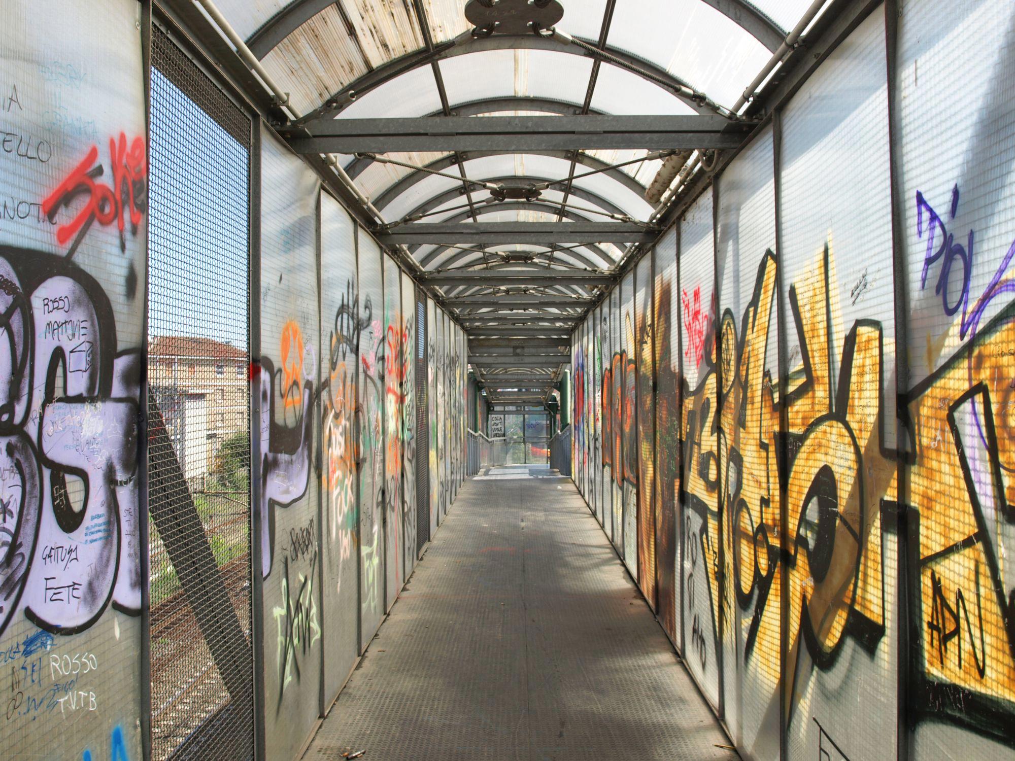 Graffiti-covered pedestrian walkway with metal mesh walls and a curved roof, highlighting urban space and the importance of community safety and upkeep.