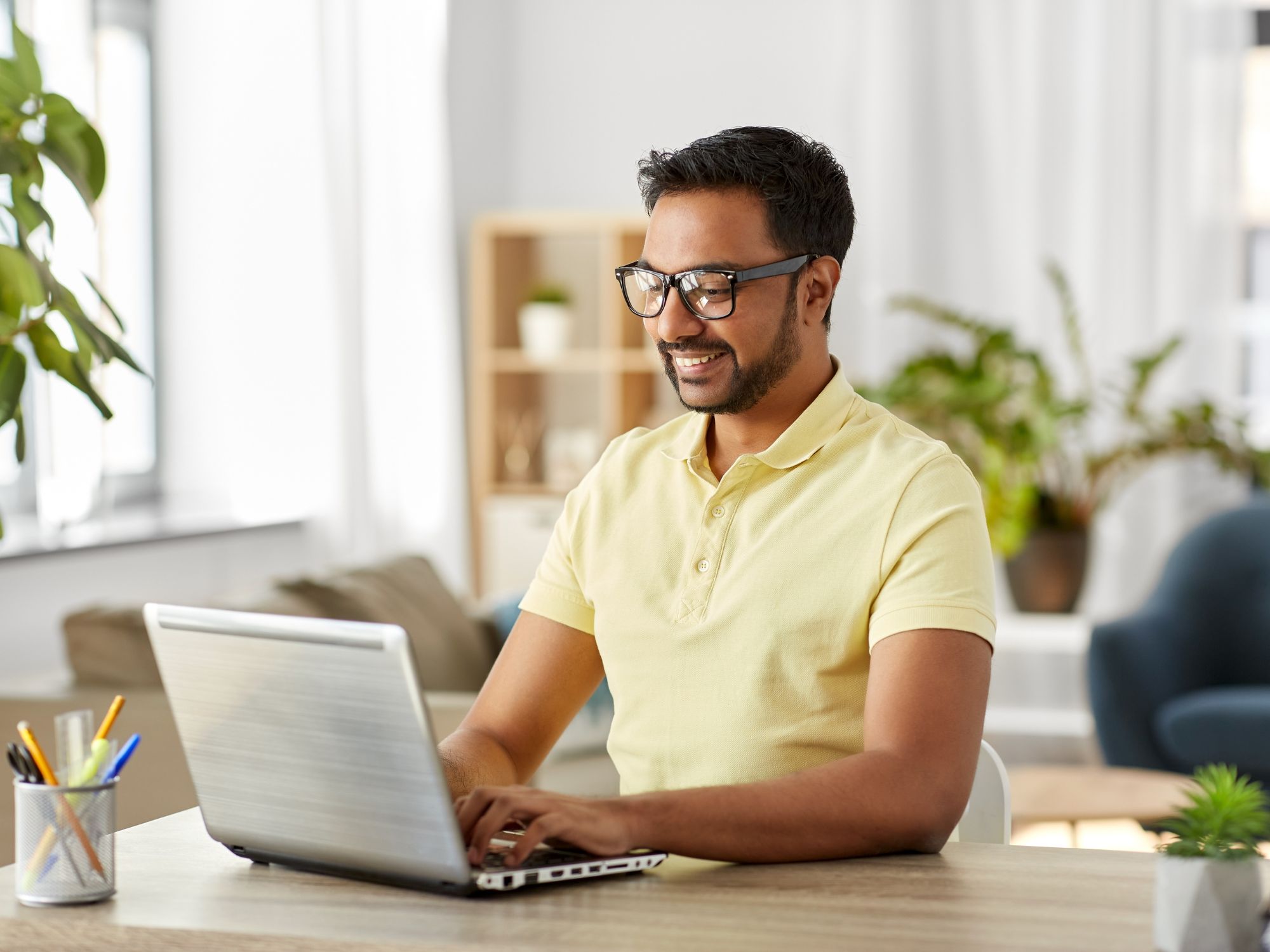 A person in a yellow polo shirt sits at a desk working on a laptop in a bright, well-decorated room with plants and shelves.