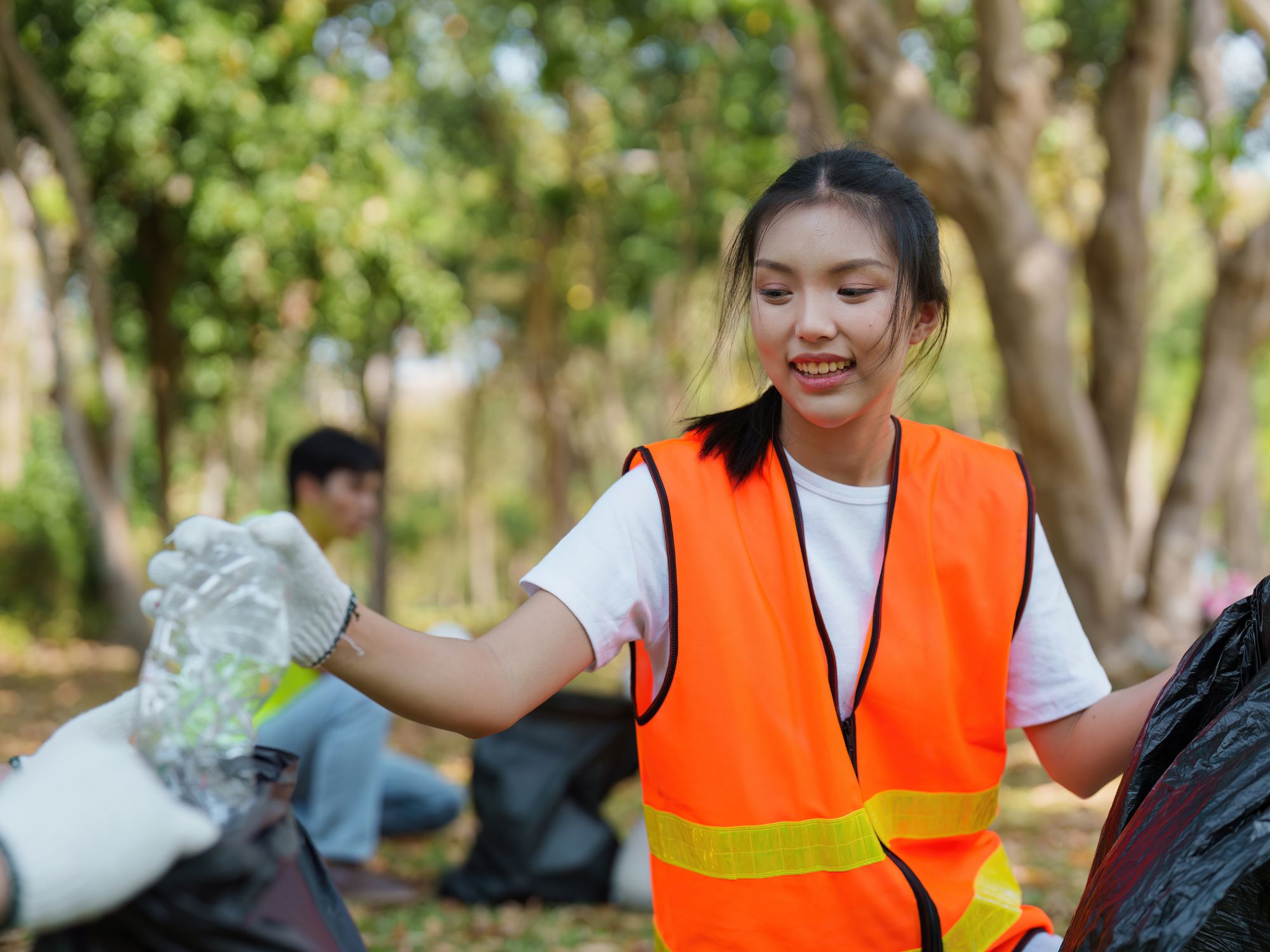 Person in an orange reflective vest and white gloves collecting litter in a wooded park area, reaching for a plastic bottle handed by another volunteer, with trees and a second participant visible in the background.