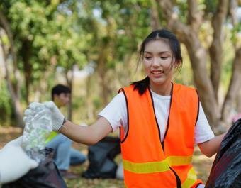 Person in an orange reflective vest and white gloves collecting litter in a wooded park area, reaching for a plastic bottle handed by another volunteer, with trees and a second participant visible in the background.