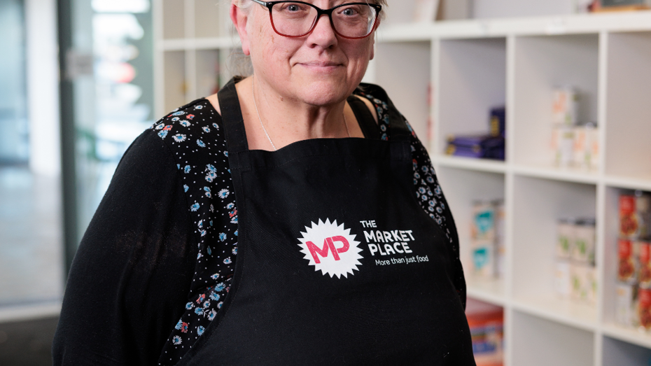Person wearing glasses and a black apron with 'MP The Market Place – Where Your Food is Free' printed on it, standing in front of shelves filled with food items.