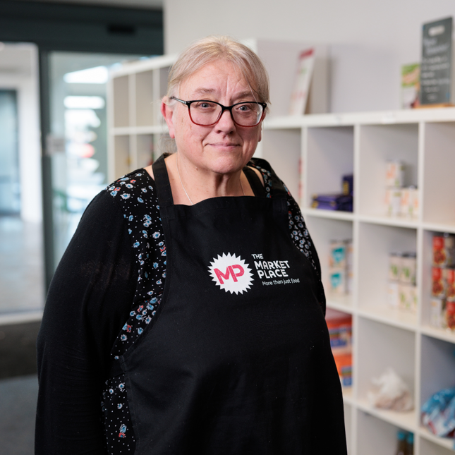 Person wearing glasses and a black apron with 'MP The Market Place – Where Your Food is Free' printed on it, standing in front of shelves filled with food items.