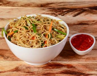 “A white bowl filled with vegetable stir-fried noodles, served on a wooden surface with a small dish of red sauce on the side.”