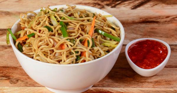 “A white bowl filled with vegetable stir-fried noodles, served on a wooden surface with a small dish of red sauce on the side.”