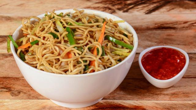 “A white bowl filled with vegetable stir-fried noodles, served on a wooden surface with a small dish of red sauce on the side.”