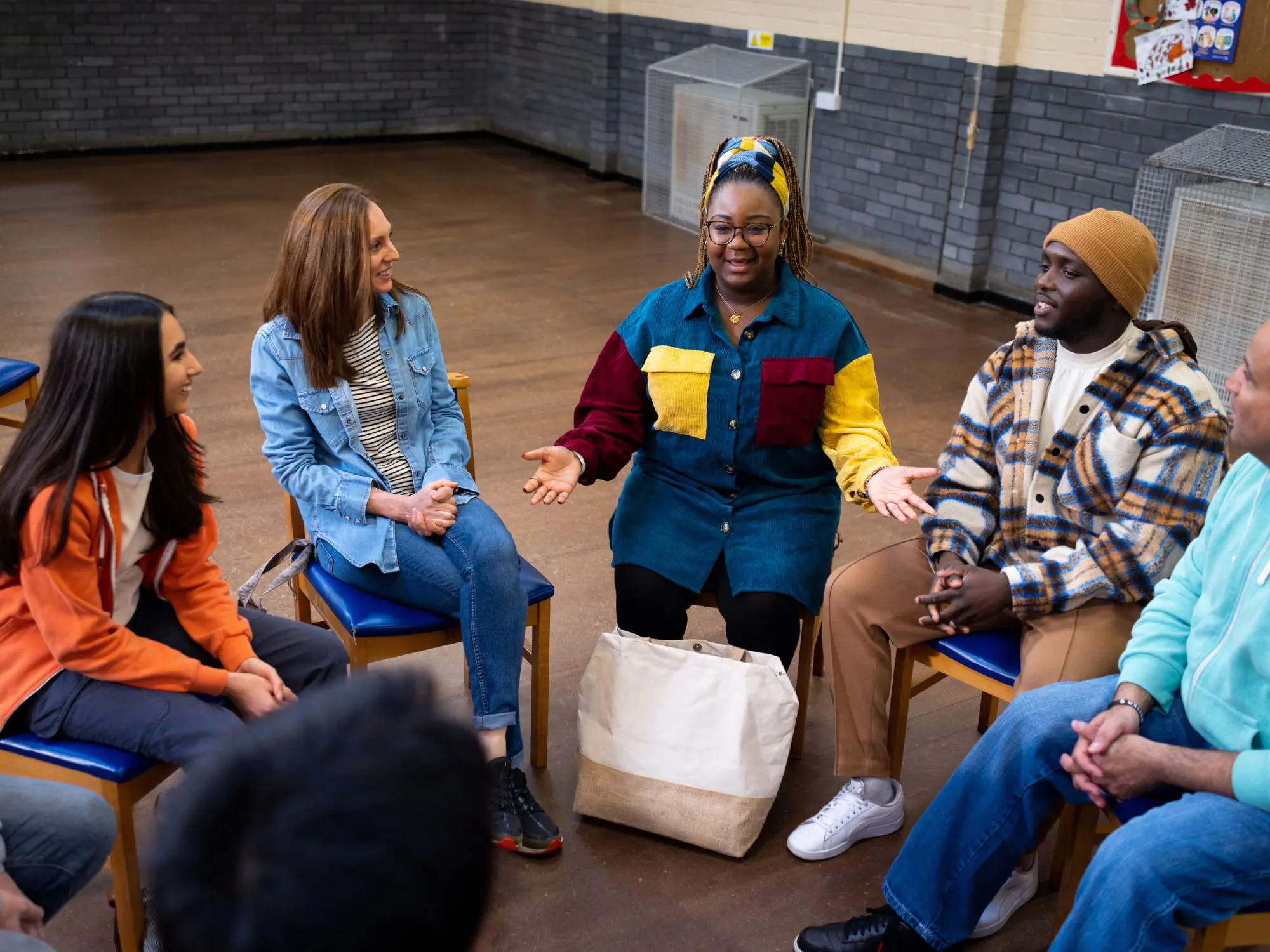 A group of people seated in a circle on blue chairs in a community hall with brick walls and a wooden floor. One person in the center, wearing a colorful patchwork shirt and headscarf, is speaking with hands open in a gesture, while others listen attentively. A large beige tote bag rests on the floor in front of the speaker.