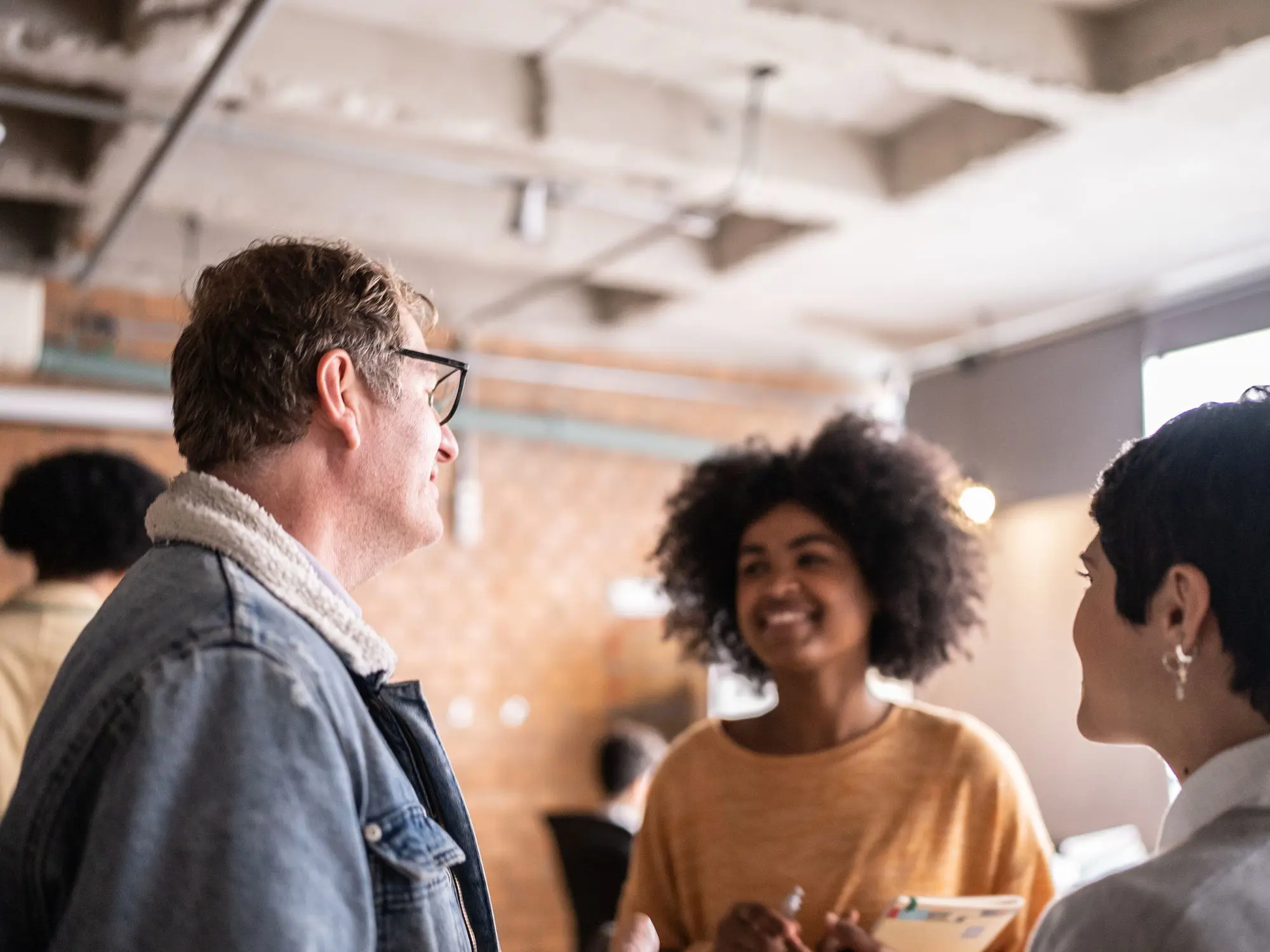 “Three people engaged in conversation indoors, holding papers in a modern space with exposed ceiling and brick walls.”