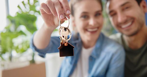 Two people sit closely together indoors, with one holding up a set of keys attached to a house-shaped keychain. Green plants and a softly blurred background suggest a warm, homey environment.