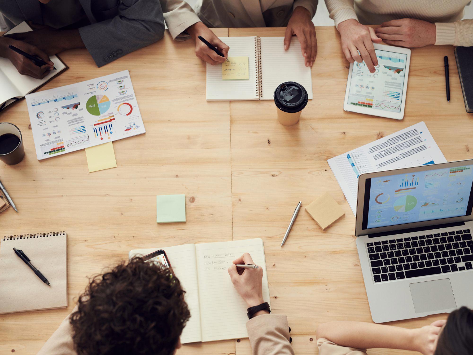 Group of people seated around a wooden table during a collaborative meeting, with notebooks, pens, sticky notes, a coffee cup, and electronic devices including a tablet and a laptop displaying charts and graphs