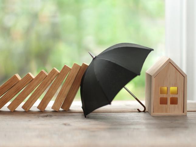 Row of falling wooden dominoes approaching a small black umbrella shielding a miniature wooden house with lit windows, set against a blurred green outdoor background. Row of falling wooden dominoes approaching a small black umbrella shielding a miniature wooden house with lit windows, set against a blurred green outdoor background.