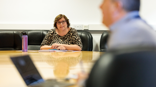 A person wearing glasses and a leopard print top sits at a conference table with a purple water bottle, papers, and notebooks, suggesting a meeting or discussion setting.
