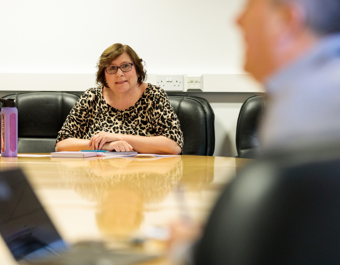 A person wearing glasses and a leopard print top sits at a conference table with a purple water bottle, papers, and notebooks, suggesting a meeting or discussion setting.