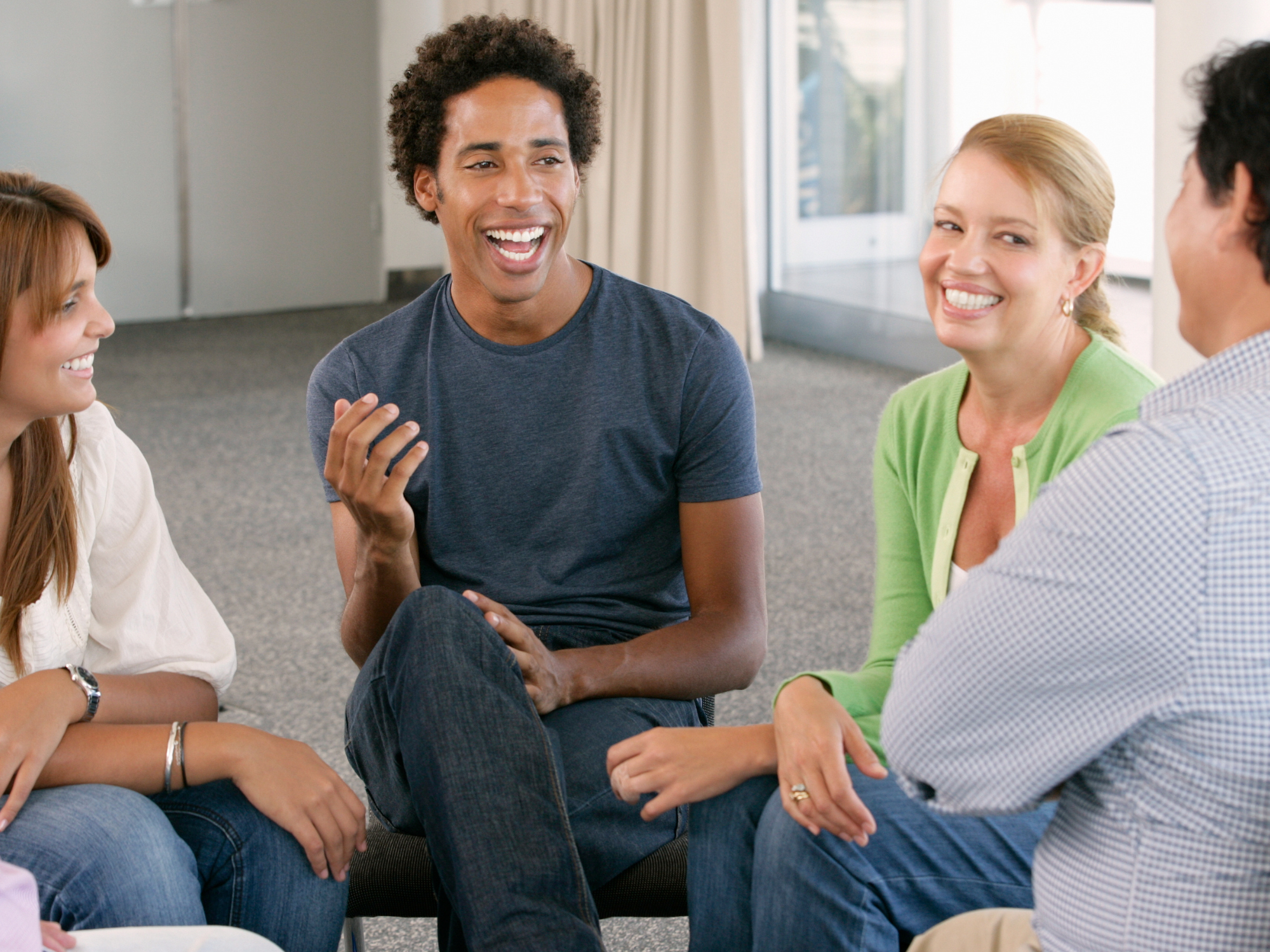 Four adults seated in a circle indoors, facing each other and engaged in conversation. One person in a dark T‑shirt gestures with a hand while others sit nearby, all positioned closely together in a bright room with windows and light-coloured walls.