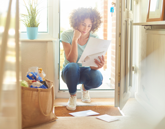 Person crouching at the entrance of a home, reading a piece of paper with several sheets in hand. A grocery bag and scattered papers are on the floor, with a potted plant visible on a windowsill in the background.