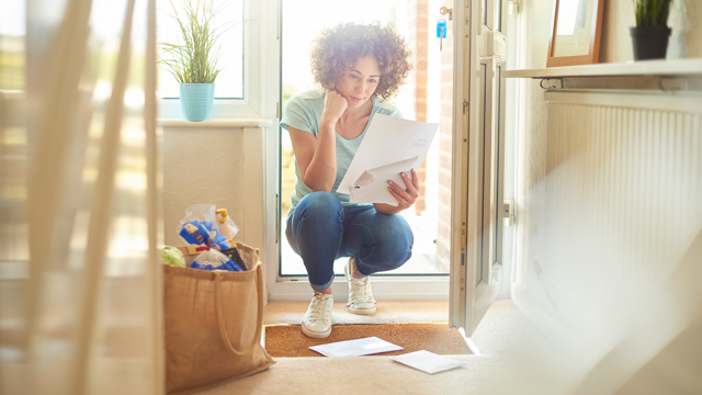 Person crouching at the entrance of a home, reading a piece of paper with several sheets in hand. A grocery bag and scattered papers are on the floor, with a potted plant visible on a windowsill in the background.