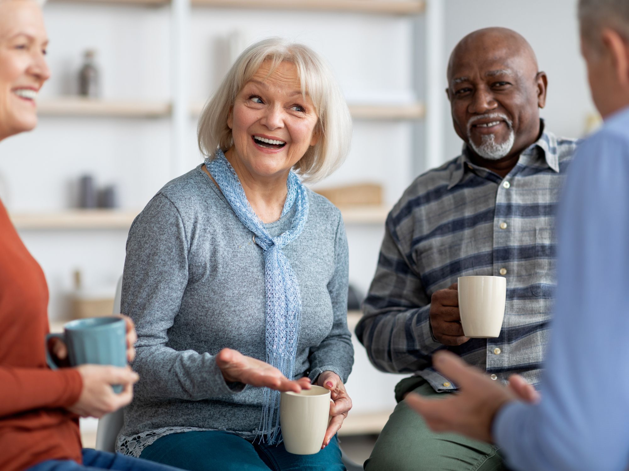 Four people sitting in a casual indoor setting, each holding a mug. They are engaged in conversation, with one person gesturing while speaking. The background shows shelves with decorative items and a light-colored wall.