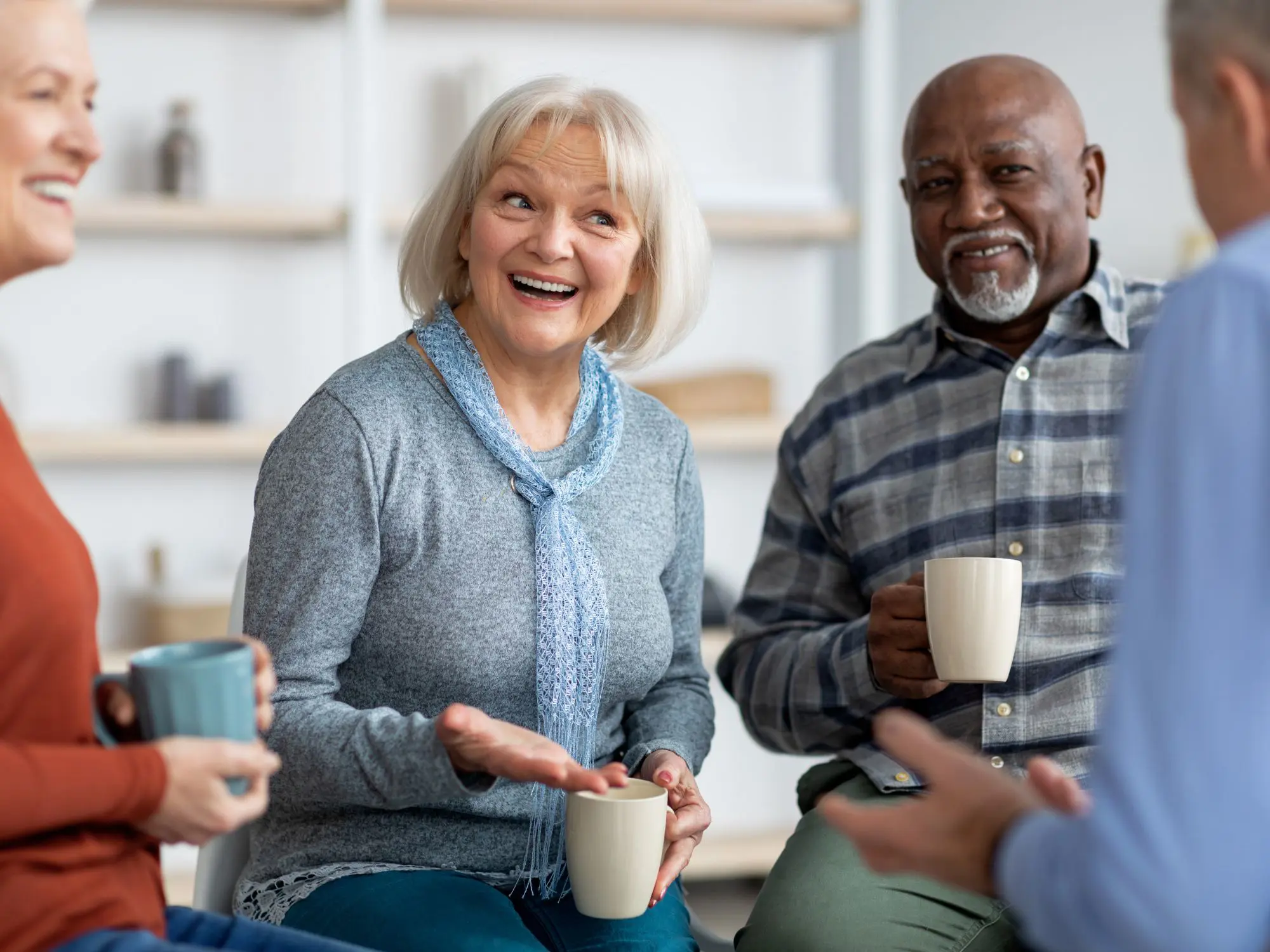 Four people sitting in a casual indoor setting, each holding a mug. They are engaged in conversation, with one person gesturing while speaking. The background shows shelves with decorative items and a light-colored wall.