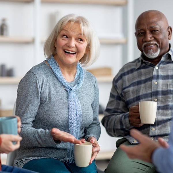 Four people sitting in a casual indoor setting, each holding a mug. They are engaged in conversation, with one person gesturing while speaking. The background shows shelves with decorative items and a light-colored wall.