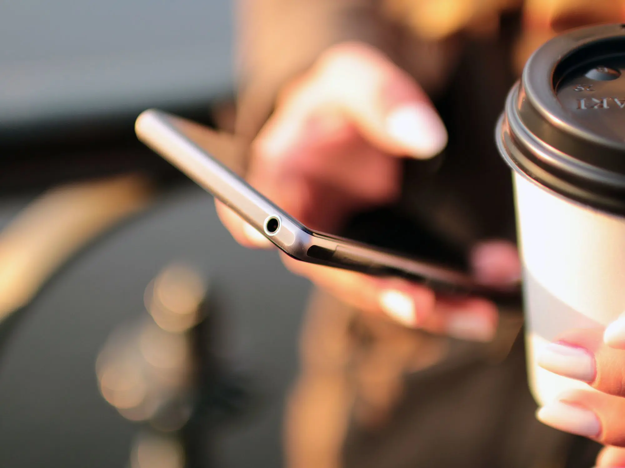 Close-up of a person holding a smartphone and a disposable coffee cup with a lid.