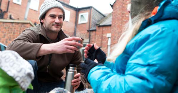 Two people interacting outdoors in a residential area. One, wearing a brown jacket and knit hat, holds a thermos and offers something to the other, who has long hair and wears a blue jacket, gloves, and a hat. Brick buildings with windows are visible in the background.