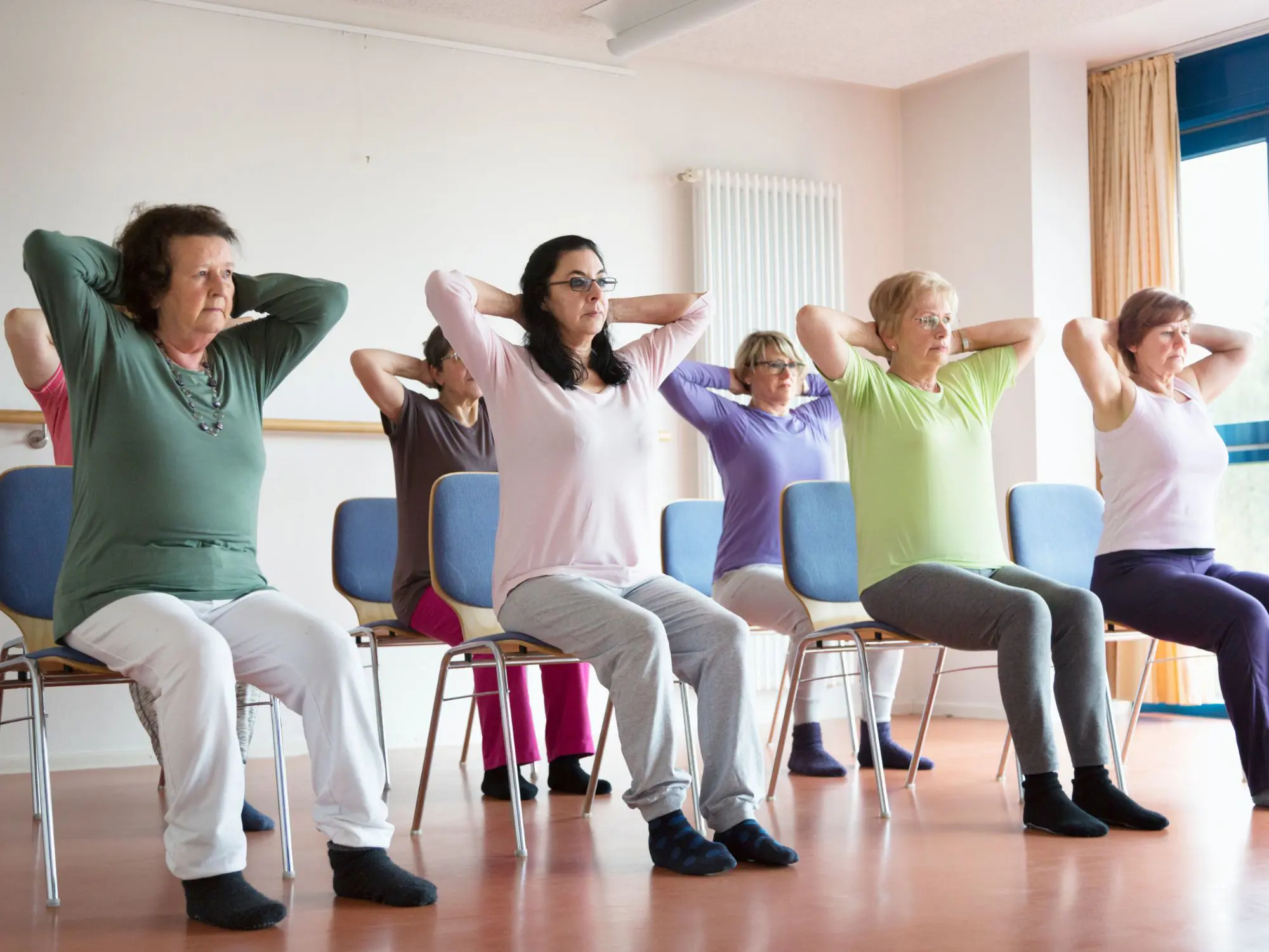 A group of people seated on chairs in a bright room performing a fitness routine. Each person has their hands placed behind their head, elbows out, and feet flat on the floor, suggesting a chair-based exercise or stretching session. The room has wooden flooring, blue cushioned chairs, a radiator on the wall, and large windows letting in natural light.
