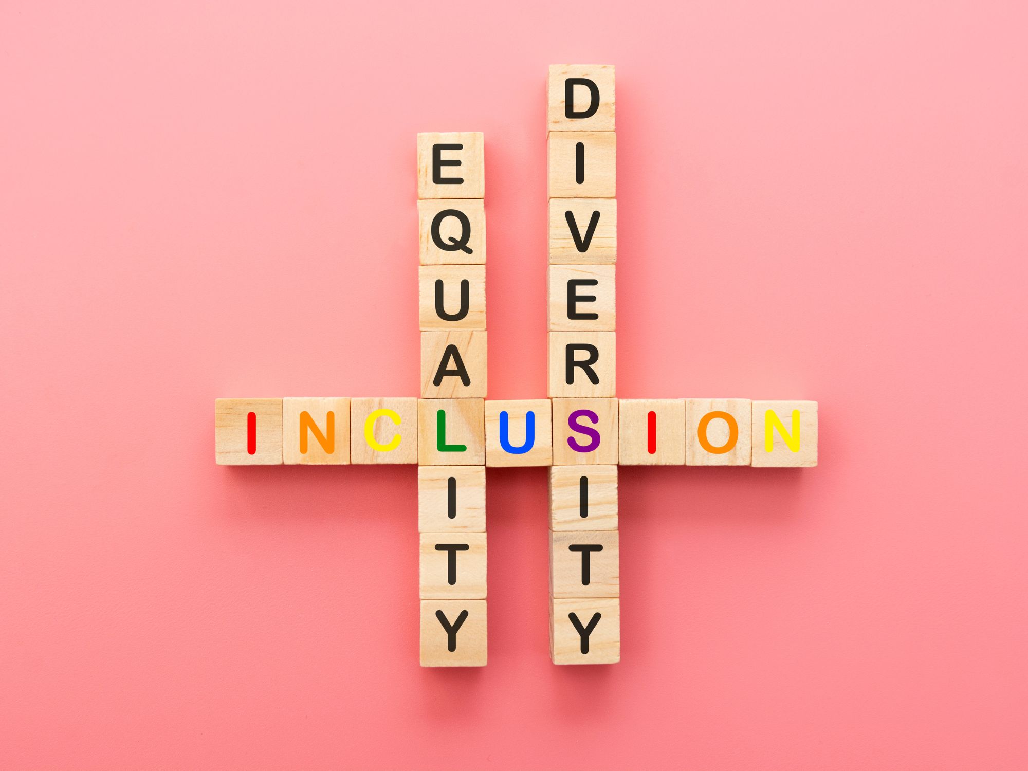 Wooden blocks arranged on a pink background spell out "EQUALITY" and "DIVERSITY" vertically, intersecting with "INCLUSION" horizontally. The letters of "INCLUSION" are in rainbow colors, while "EQUALITY" and "DIVERSITY" are in black, representing the interconnectedness of equality, diversity, and inclusion.