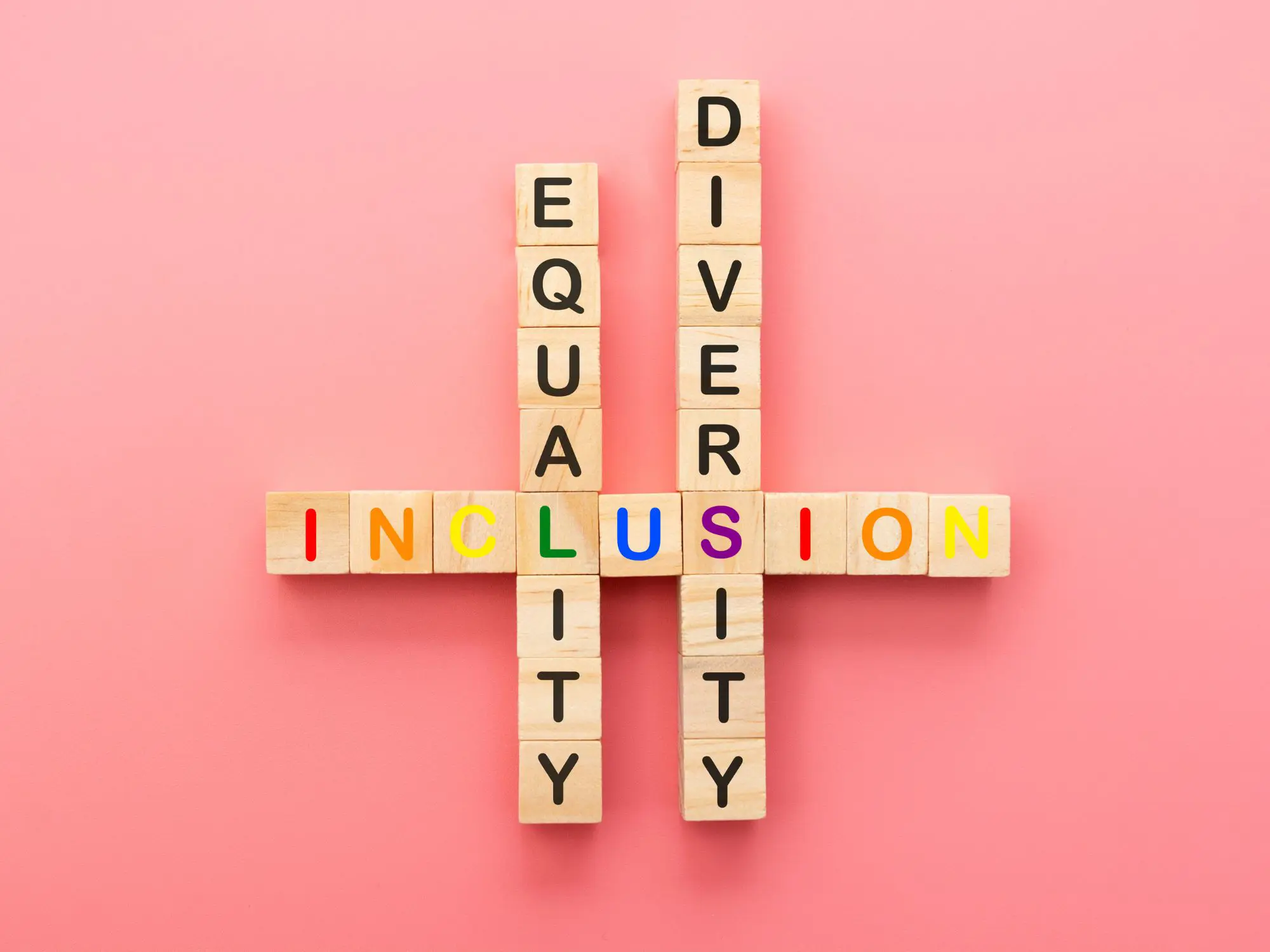 Wooden blocks arranged on a pink background spell out "EQUALITY" and "DIVERSITY" vertically, intersecting with "INCLUSION" horizontally. The letters of "INCLUSION" are in rainbow colors, while "EQUALITY" and "DIVERSITY" are in black, representing the interconnectedness of equality, diversity, and inclusion.