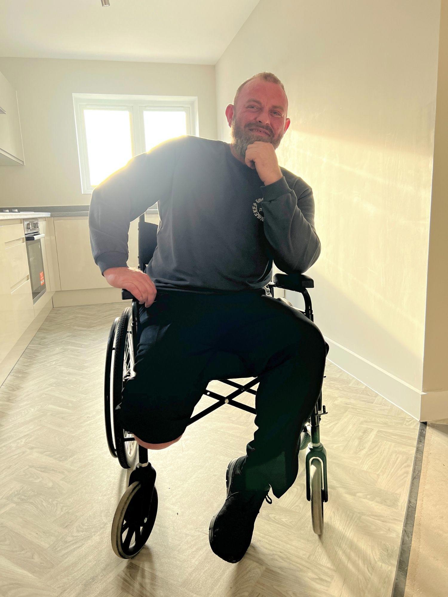 A man sitting in a wheelchair in a kitchen, smiling at the camera, backlit with warm sunshine through the kitchen window.
