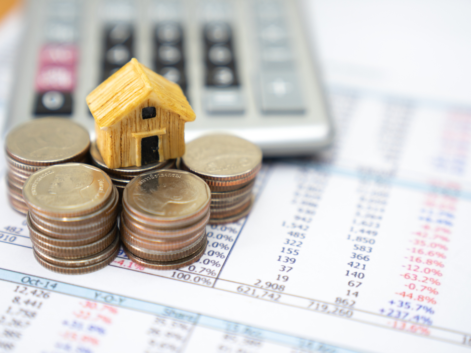 Small wooden house model sitting on stacks of coins, with a calculator and financial figures in the background.