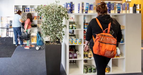 Three people browsing shelves stocked with food items at The Market Place; one wears an orange backpack, and a large potted plant sits in front of the shelves in a carpeted, fluorescent-lit room.