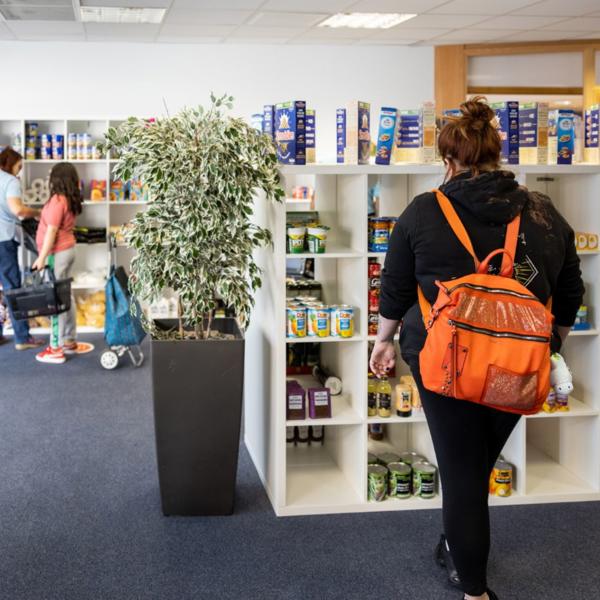 Three people browsing shelves stocked with food items at The Market Place; one wears an orange backpack, and a large potted plant sits in front of the shelves in a carpeted, fluorescent-lit room.