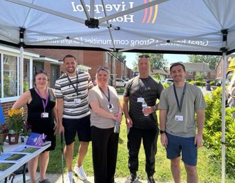 A group of five staff wearing lanyards pose under a South Liverpool Homes gazebo at a local community event, with an information table, plants and a row of houses visible behind.
