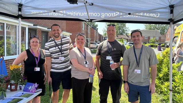 A group of five staff wearing lanyards pose under a South Liverpool Homes gazebo at a local community event, with an information table, plants and a row of houses visible behind.