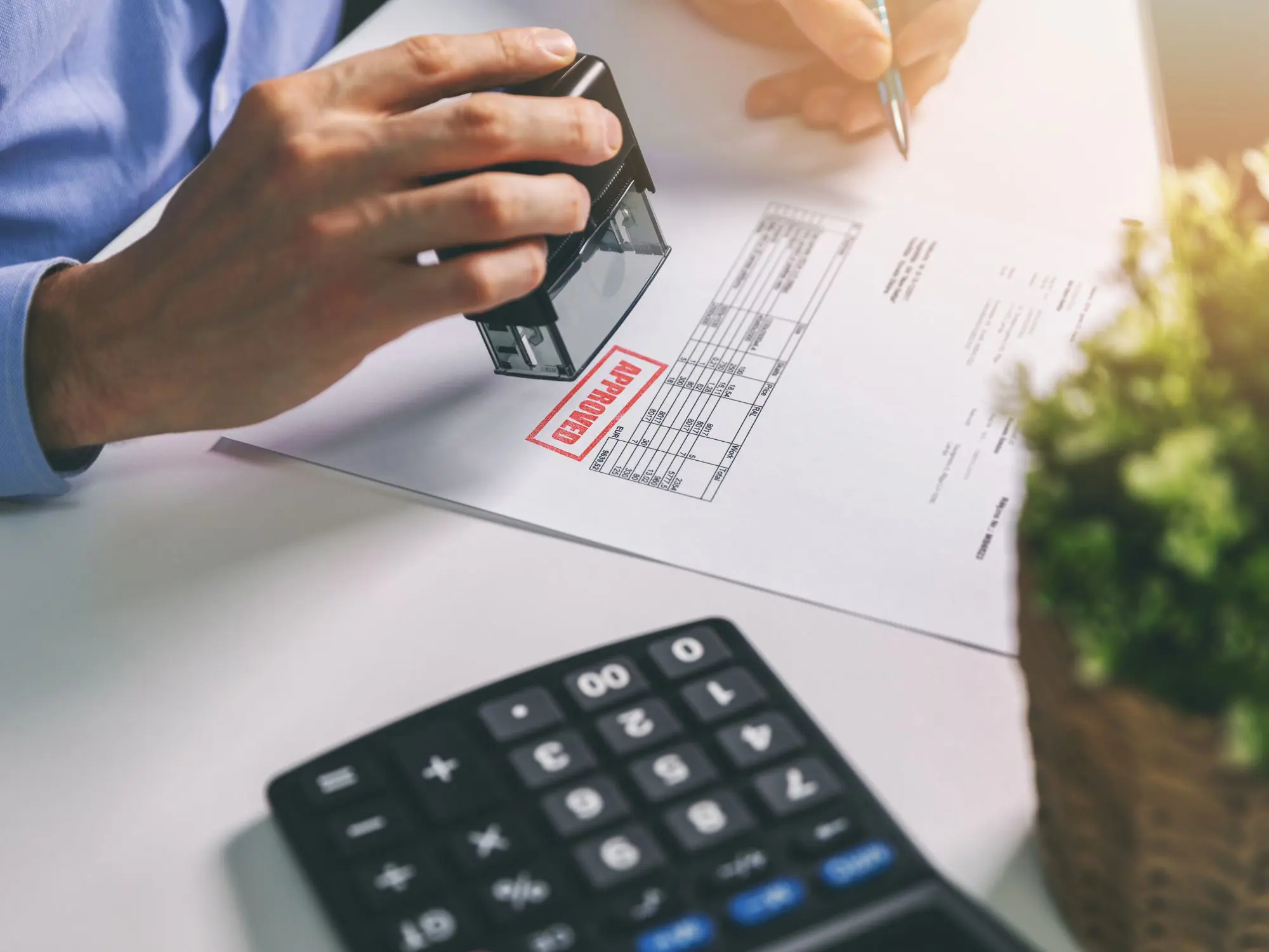 A person stamps a document marked 'APPROVED' beside a calculator and a potted plant, suggesting formal approval of a report or procurement paperwork.