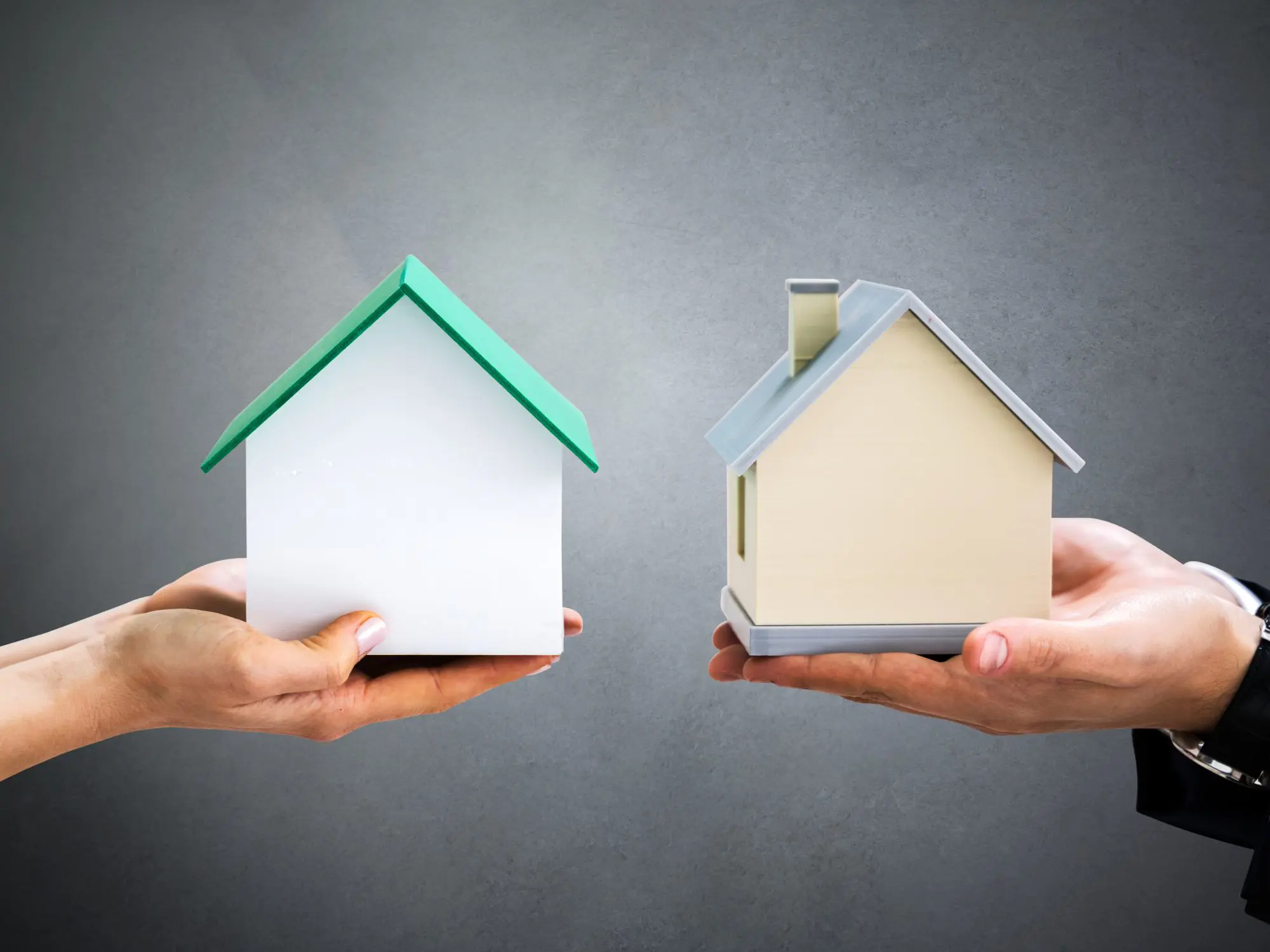 Two hands holding small house models against a plain gray background—one white with a green roof, the other beige with a gray roof and chimney.