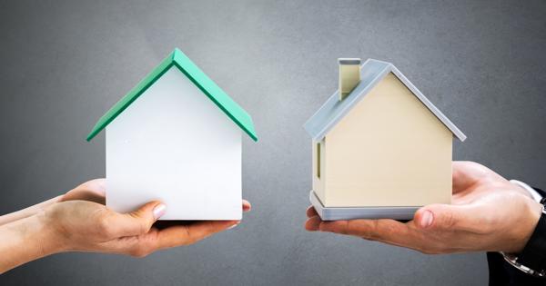 Two hands holding small house models against a plain gray background—one white with a green roof, the other beige with a gray roof and chimney.